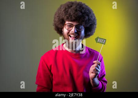 Fröhlicher positiver Hipster-Mann mit Afro-Frisur, der eine Veranstaltung veranstaltet, Party-Requisiten mit einem schönen Wort in den Händen hält und ein rotes Sweatshirt trägt. Innenaufnahmen im Studio, isoliert auf farbigem Neonlicht-Hintergrund. Stockfoto