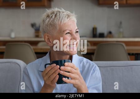 Glückliche ältere Frau sitzt auf dem Sofa hält den Becher und schaut zur Seite Stockfoto