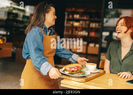 Glückliche Kellnerin mit Down-Syndrom serviert einem Gast ein Sandwich und Kaffee in einem trendigen Café. Professionelle Frau mit einer intellektuellen Behinderung arbeitet Stockfoto