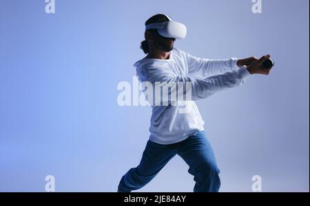Junger Mann, der Kricket in der virtuellen Realität spielt. Sportlicher junger Mann, der mit Gaming-Controllern einen virtuellen Ball schlägt. Aktiver junger Mann, der immersive V erforscht Stockfoto