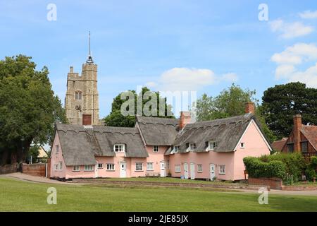 Strohgedeckte Häuschen, pink mit der Kirche der Heiligen Jungfrau Maria auf dem Dorf grün gestrichen. Cavendish, Suffolk, East Anglia, Großbritannien Stockfoto