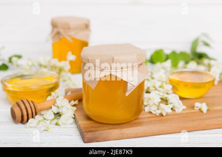 Süßes Honigglas umgab die Akazienblüten des Frühlings. Honig fließt aus einem Löffel in ein Glas. Gläser von klarem frischen Akazienhonig auf Holzgrund. Stockfoto