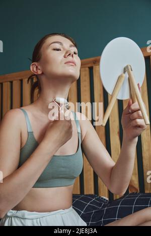 Junge Frau mit Jade Gesichtswalze für Gesichtsmassage sitzen auf dem Bett im Schlafzimmer Blick in den Spiegel Stockfoto