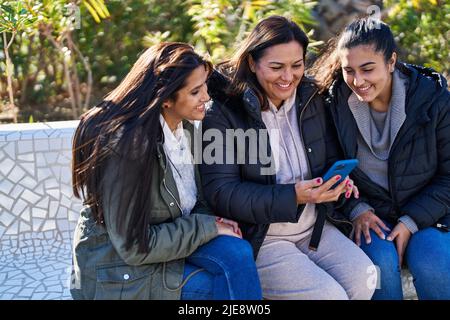 Drei weibliche Mutter und Töchter mit Smartphone sitzen auf der Bank im Park Stockfoto