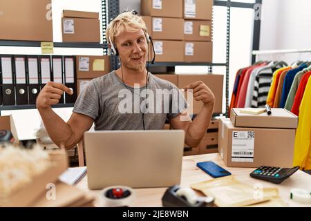 Der junge blonde Mann, der ein Headset trägt und im Online-Shop arbeitet, sieht selbstbewusst mit einem Lächeln im Gesicht aus und zeigt sich mit stolzen und glücklichen Fingern. Stockfoto