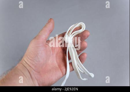 Ein Mann, der eine weiße Trageschlaufe in der Hand hält. Grauer Hintergrund. Flacher Schnürsenkel mit Knoten und Schlaufe liegt in offener Handfläche. Reifer Mann. Nahaufnahme. Selektiver Fokus. Stockfoto