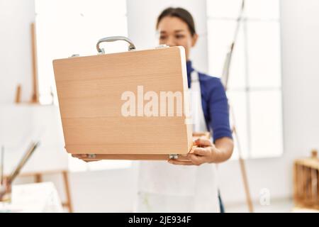 Junge latinerin lächelt selbstbewusst und eröffnet eine Aktentasche im Kunststudio Stockfoto