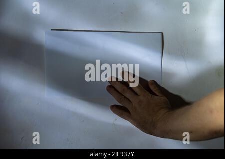 Die Hand eines reifen Mannes drückt ein Blatt Papier gegen die Wand. Leeres weißes rechteckiges Stück Papier, das an der Wand befestigt ist. Abstraktes Licht und Schatten zeichnen sich ab Stockfoto