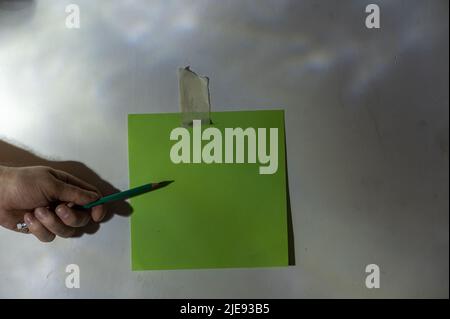 Die Hand des reifen Mannes zeigt mit Bleistift auf ein Stück Papier. Leeres grünes quadratisches Stück Papier, das an der Wand befestigt ist. Eine abstrakte Licht- und Schattenzeichnung auf einem Stockfoto