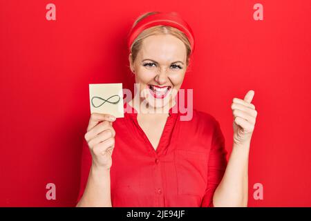 Junge blonde Frau hält unendliche Symbol Erinnerung schreiend stolz, feiert Sieg und Erfolg sehr aufgeregt mit erhobenen Armen Stockfoto