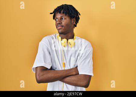 Junger afrikanischer Mann mit Dreadlocks, die auf gelbem Hintergrund stehen und mit überkreuzten Armen zur Seite schauen, überzeugt und zuversichtlich Stockfoto