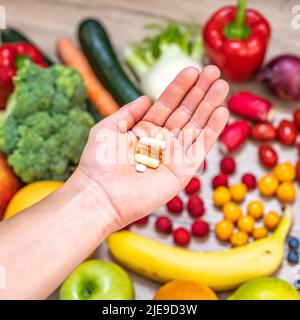 Hand hält Nahrungsergänzungstabletten über einem Holztisch voller Gemüse und Obst. Nahrungsergänzungsmittel ermöglichen einen gesunden Lebensstil. Stockfoto