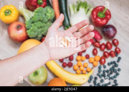 Hand hält Nahrungsergänzungstabletten über einem Holztisch voller Gemüse und Obst. Nahrungsergänzungsmittel ermöglichen einen gesunden Lebensstil. Stockfoto