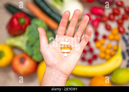 Hand hält Nahrungsergänzungstabletten über einem Holztisch voller Gemüse und Obst. Nahrungsergänzungsmittel ermöglichen einen gesunden Lebensstil. Stockfoto