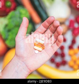 Hand hält Nahrungsergänzungstabletten über einem Holztisch voller Gemüse und Obst. Nahrungsergänzungsmittel ermöglichen einen gesunden Lebensstil. Stockfoto