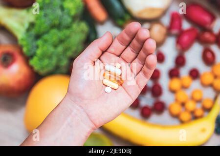 Hand hält Nahrungsergänzungstabletten über einem Holztisch voller Gemüse und Obst. Nahrungsergänzungsmittel ermöglichen einen gesunden Lebensstil. Stockfoto