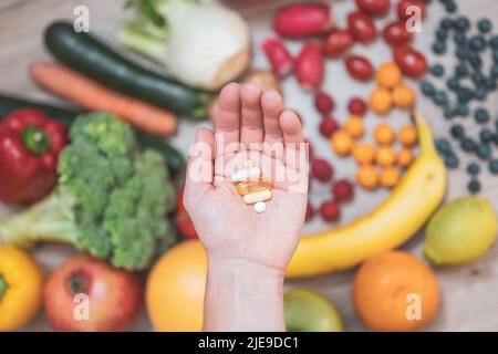 Hand hält Nahrungsergänzungstabletten über einem Holztisch voller Gemüse und Obst. Nahrungsergänzungsmittel ermöglichen einen gesunden Lebensstil. Stockfoto