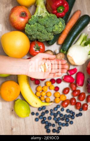 Hand hält Nahrungsergänzungstabletten über einem Holztisch voller Gemüse und Obst. Nahrungsergänzungsmittel ermöglichen einen gesunden Lebensstil. Stockfoto