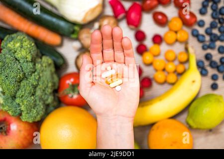 Hand hält Nahrungsergänzungstabletten über einem Holztisch voller Gemüse und Obst. Nahrungsergänzungsmittel ermöglichen einen gesunden Lebensstil. Stockfoto