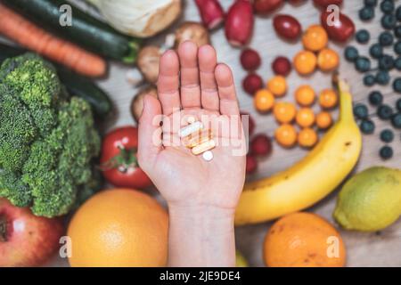 Hand hält Nahrungsergänzungstabletten über einem Holztisch voller Gemüse und Obst. Nahrungsergänzungsmittel ermöglichen einen gesunden Lebensstil. Stockfoto