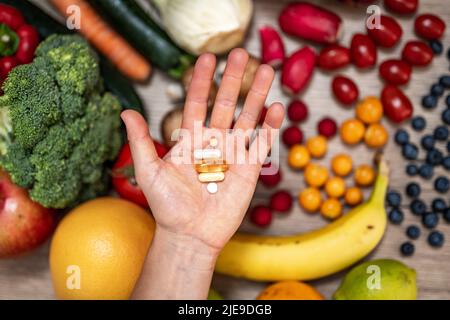 Hand hält Nahrungsergänzungstabletten über einem Holztisch voller Gemüse und Obst. Nahrungsergänzungsmittel ermöglichen einen gesunden Lebensstil. Stockfoto