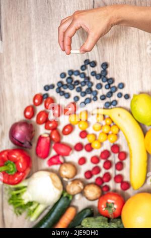 Hand hält Nahrungsergänzungstabletten über einem Holztisch voller Gemüse und Obst. Nahrungsergänzungsmittel ermöglichen einen gesunden Lebensstil. Stockfoto