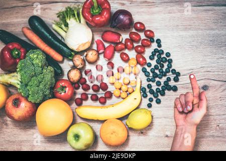 Hand hält Nahrungsergänzungstabletten über einem Holztisch voller Gemüse und Obst. Nahrungsergänzungsmittel ermöglichen einen gesunden Lebensstil. Stockfoto