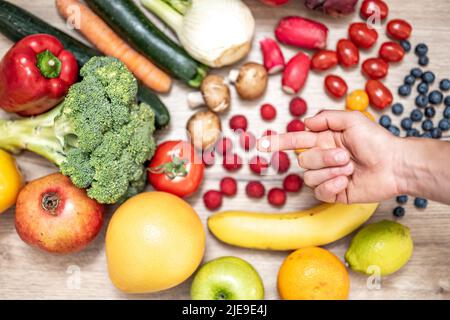 Hand hält Nahrungsergänzungstabletten über einem Holztisch voller Gemüse und Obst. Nahrungsergänzungsmittel ermöglichen einen gesunden Lebensstil. Stockfoto