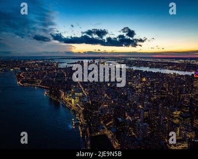 Atemberaubende Panoramaaufnahme der Stadt, die vom Fluss umgeben ist. Beleuchtete Fenster und farbenfroher Sonnenuntergang. Manhattan, New York City, USA Stockfoto