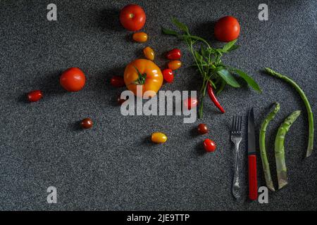 Lebensmittelrand mit grünem Spargel, roten und orangen Tomaten und rotem Chili mit Blättern. Gemüse, rotes Messer und Gabel auf dunkelgrauem Hintergrund t Stockfoto