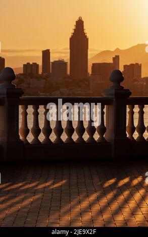 Blick auf die Stadt Benidorm in der untergehenden Sonne, Sonnenuntergang, selektiver Fokus auf den Schatten der Balustrade im Vordergrund. Stockfoto