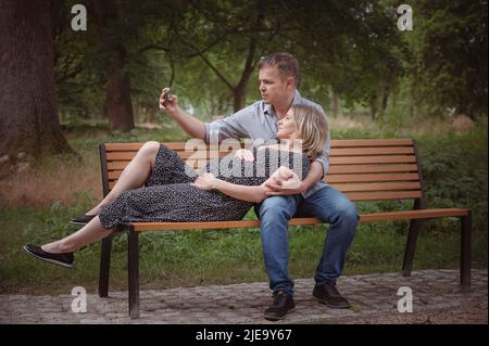 Schwanger Frau mit Mann auf Bank. Mann, der im öffentlichen Park Selfie mit seiner Frau über das Mobiltelefon gemacht hat. Liebevolles Paar genießen - warten Geburt des Babys Stockfoto