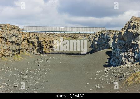 Metallbrücke über den Boden auf der isländischen Halbinsel Reykjanes. Schwarzer Lavasand mit kleinen Steinen am Boden. Geographische Risse im nordamerikanischen an Stockfoto