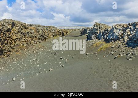 Schwarzer Lavasand mit kleinen Steinen am Boden. Metallbrücke auf Island auf der Halbinsel Reykjanes. Geographische Risse der nordamerikanischen und eurasischen Länder Stockfoto