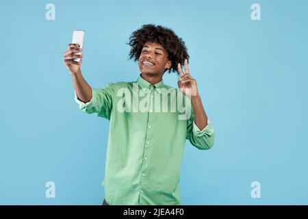 Aufgeregter afroamerikanischer Kerl, der Selfie auf dem Smartphone macht und V-Zeichen zeigt, live im Web streamt, blauer Hintergrund Stockfoto