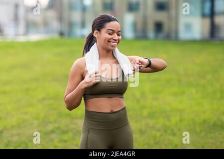 Porträt einer sportlichen jungen schwarzen Frau mit einem Handtuch, das ihre Smartwatch oder ihren Fitnesstracker im Stadtpark überprüft Stockfoto