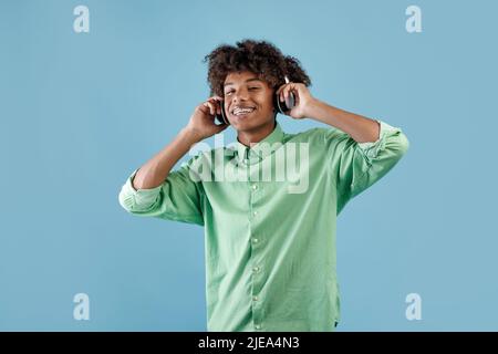 Glücklicher, friedlicher afroamerikanischer Kerl, der Musik in einem schnurlosen Headset hört, die Augen schließt und sich auf einem entspannenden, blauen Hintergrund entspannt Stockfoto