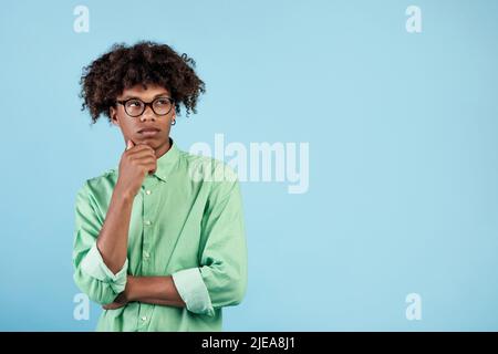 Nachdenklicher afroamerikanischer Kerl, der über etwas auf blauem Studiohintergrund nachdenkt und auf freien Raum schaut Stockfoto