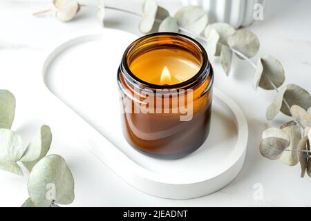 Brennende Kerze im Glas aus Bernstein auf einem trendigen Tablett aus Beton und Eukalyptuszweige auf weißem Marmorboden, Nahaufnahme. Stockfoto