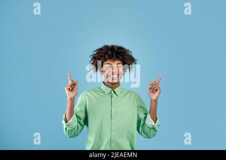 Suchen Sie nach. Glücklicher afroamerikanischer Kerl, der die Finger nach oben zeigt und lächelt auf die Kamera, zeigt etwas, blauen Hintergrund