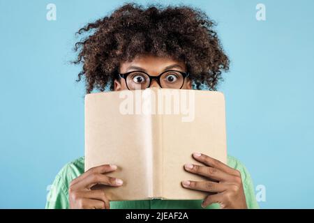 Porträt eines emotionalen afroamerikanischen Mannes in einer Brille, die das Gesicht mit einem Buch bedeckt und sich hinter ihm mit weit geöffneten Augen versteckt Stockfoto