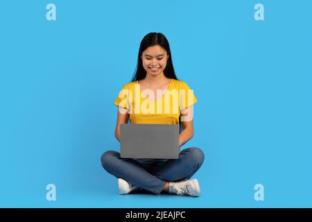Lächelnd konzentrierte junge japanische Frau digitaler Nomade in gelbem T-Shirt sitzen auf dem Boden und tippen auf dem Laptop Stockfoto