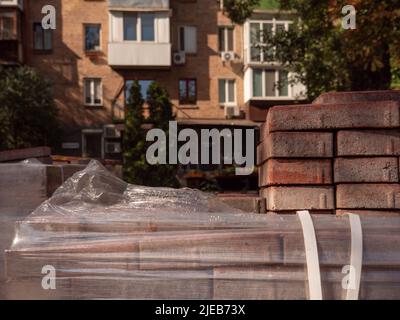 Ein Stapel Pflasterfliesen und eine Packung Zigaretten im Vordergrund und ein altes rotes Ziegelgebäude, das im Hintergrund von untergehenden Sonnenstrahlen in Kiew, Ukraine, beleuchtet wird. Stockfoto