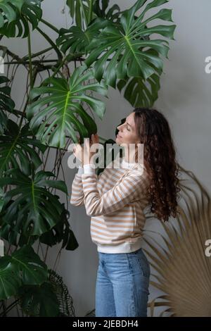 Junge lächelnde Frau berührt das Monstera-Pflanzenblatt und genießt die Gartenarbeit und plantcare Stockfoto