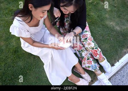 Draufsicht auf zwei Freunde, die auf dem Gras sitzen und dabei auf das Handy schauen Stockfoto