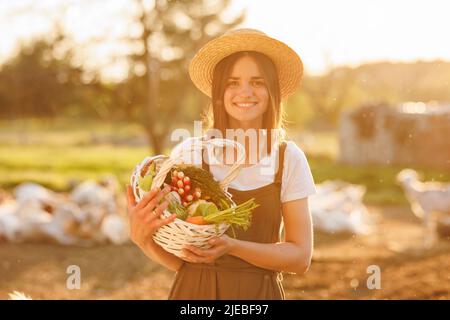 Junge kaukasische schöne Bäuerin in Strohhut mit Korb mit frischem Bio-Gemüse bei Sonnenuntergang. Vegetarische, rohe, gesunde Lebensmittel. Gesund Stockfoto