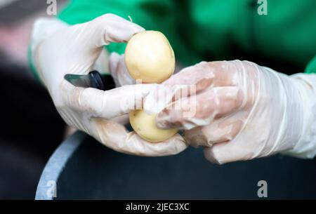 Die Hände der Frau in weißen Handschuhen schälen rohe neue Kartoffeln, Nahaufnahme. Reinigung von Bio-Kartoffeln. Detail der weiblichen Hände, die frische gelbe Kartoffel mit Kitc schälen Stockfoto