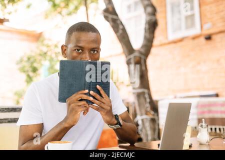 Lächelnder, fröhlicher afroamerikanischer Mann, der im Café am Tisch sitzt und seinen Mund mit einem Laptop durch ein Buch bedeckt Stockfoto