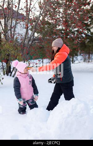 Lächelnde Mutter klebte Karotte an kleine Tochter Gesicht als Nase auf Hinterhof voller Schnee am Abend auf dem Spaziergang mit Bäumen und Eisenzaun im Hintergrund Stockfoto