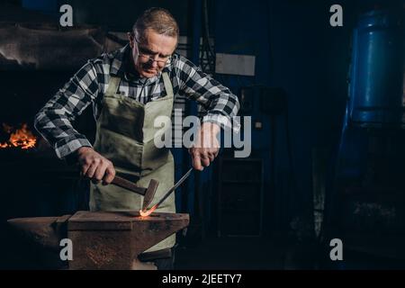 Der Schmied schmiedet das rotglühende Metall manuell auf dem Amboss in der Schmiede. Harte Arbeit. Energie und Leistung. Konzept der Arbeit, Retro-Berufe Stockfoto